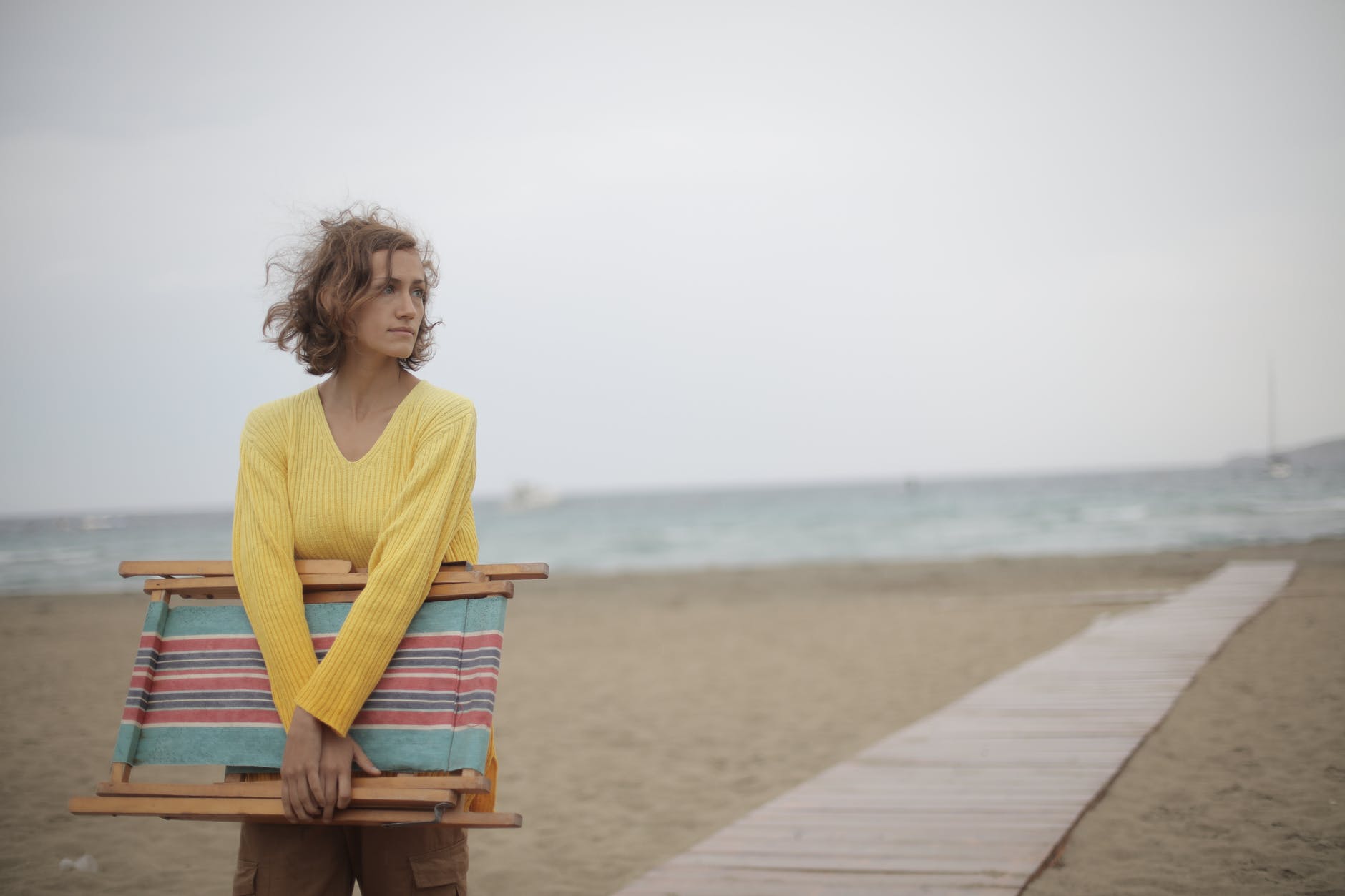 photo of woman in yellow long sleeve shirt standing at the beach carrying wooden folding beach chair body insecurity can affect everyone